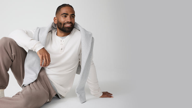 Man sitting on a white floor wearing a Vuori light gray jacket over a white shirt and beige pants.