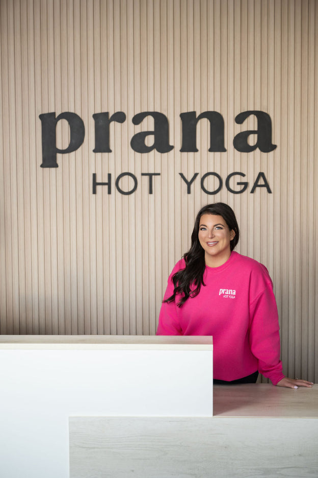 Leah Cain, founder of Prana Hot Yoga, smiling at the studio's front desk ready to answer your questions