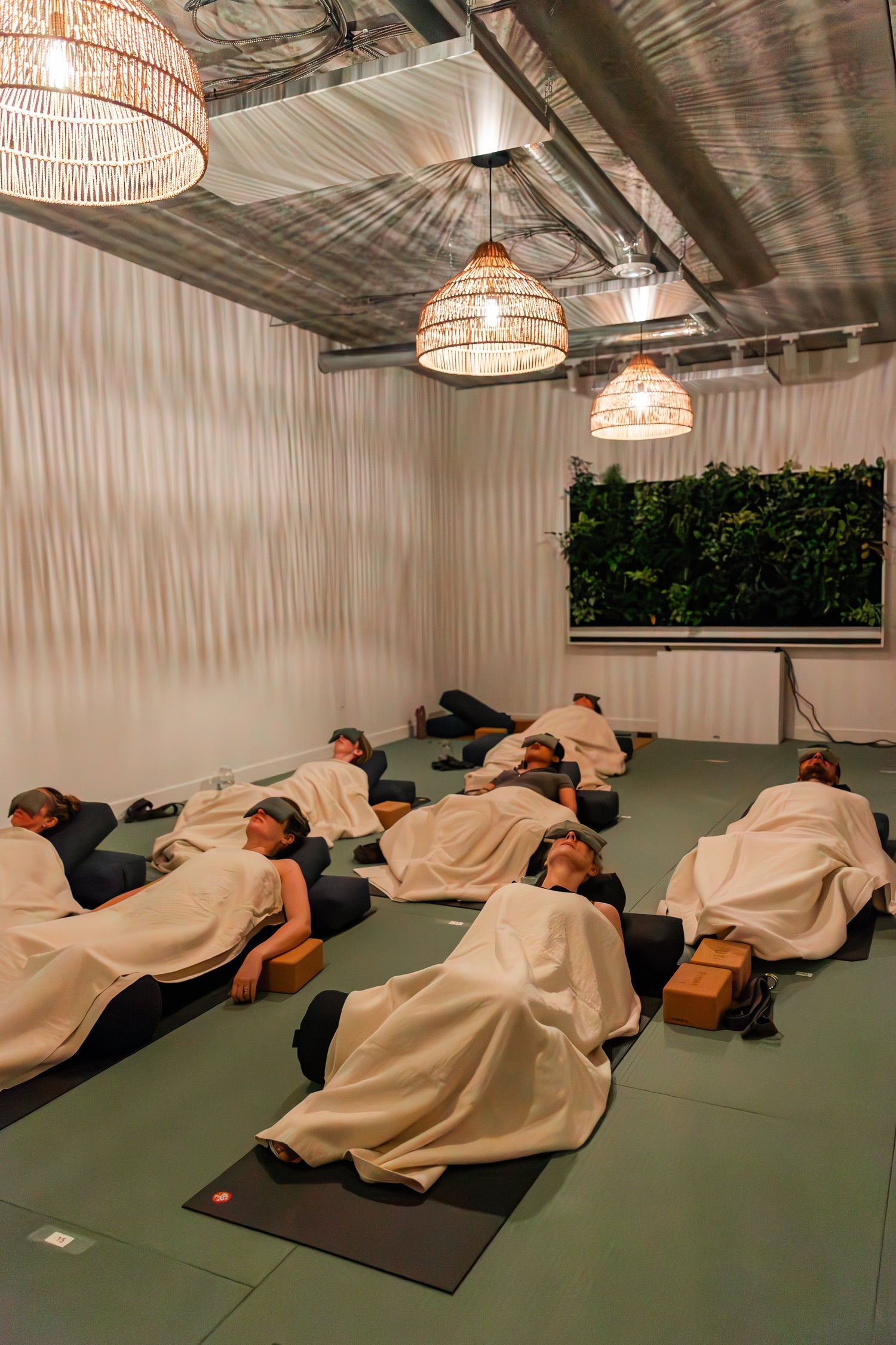 Private restorative yoga class at Prana Hot Yoga's Moon Room with participants in supported relaxation pose, blankets provided, featuring living plant wall for peaceful atmosphere